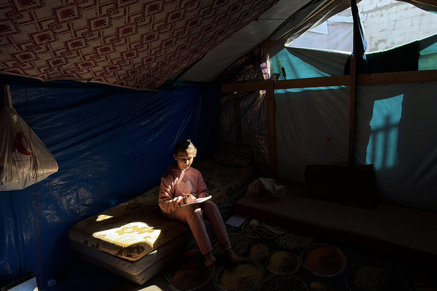 A child dressed in salmon-coloured top and pants sitting in a tent looking at a book in the dark.