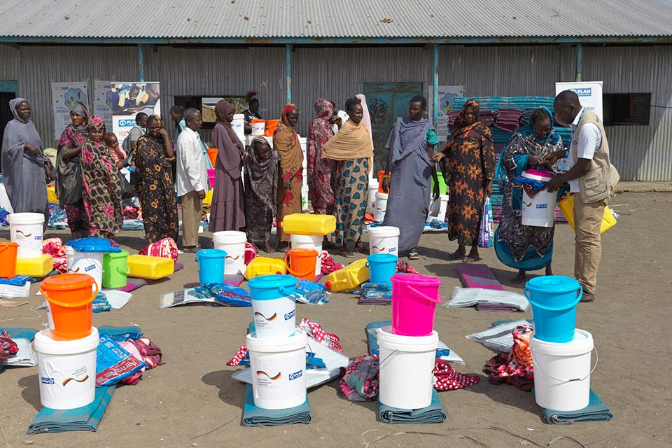 A group of people line up to collect kits that include blankets, buckets, mosquito nets, jerrycans, mats and bars of soap. 