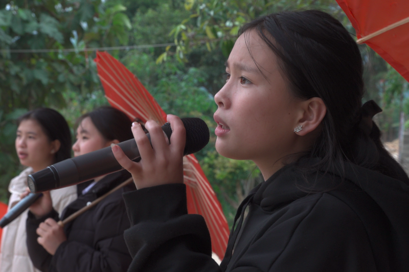 A young woman wearing black sings while holding a microphone in front of two other youth girls with red umbrellas.