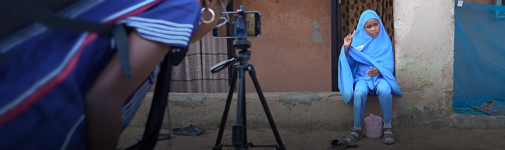 Younge female filmmaker stands behind camera filming young girl.