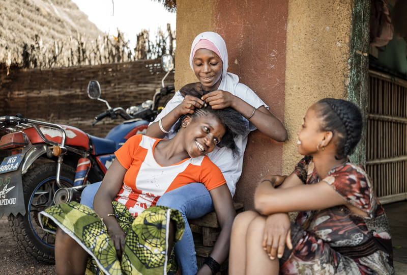 Three teenage girls sit outside, while one of the girls is braiding one girl’s hair. All three of them are smiling.