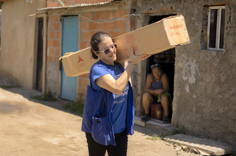 A woman in a blue Plan-branded vest carries a long box over her shoulder as she walks down a street.