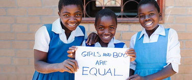 Three girls grinning and holding a "Girls and boys are equal" sign, while standing shoulder by shoulder in front of a school