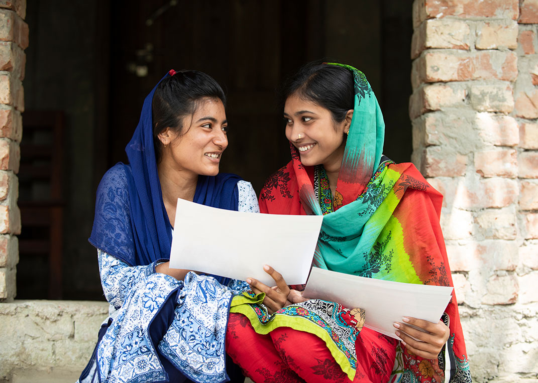 Bithi and a friend smile while sitting reading materials from Plan International
