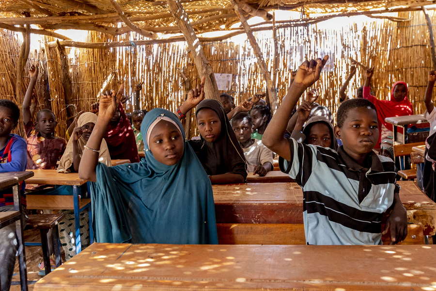 Students raise their hands behind rows of desks in an informal classroom.
