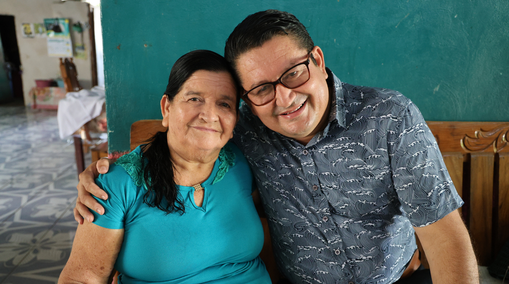Two people sit side by side indoors on a wooden bench, one wearing a turquoise shirt with dark hair, the other wearing glasses and a patterned shirt.
