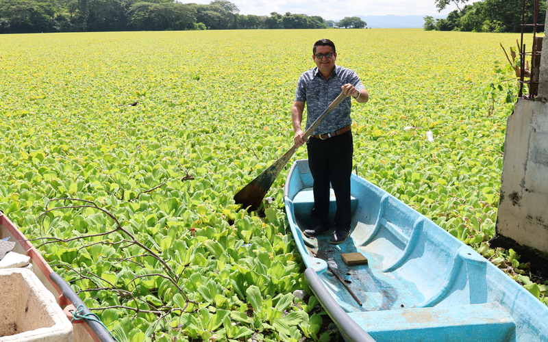 A man stands in a blue boat, paddling through a dense field of green aquatic plants. Nearby, a red and white boat holds several items. 