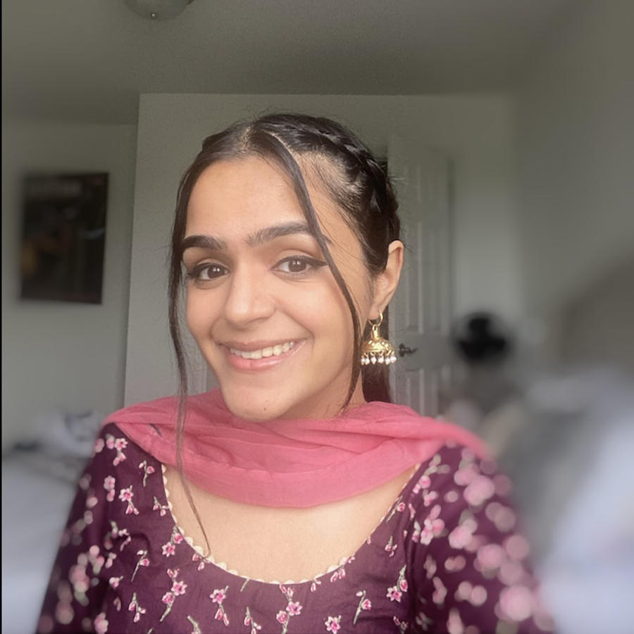 A young woman smiles at the camera. She is wearing a purple shirt with pink flowers and a pink drape around her neck.