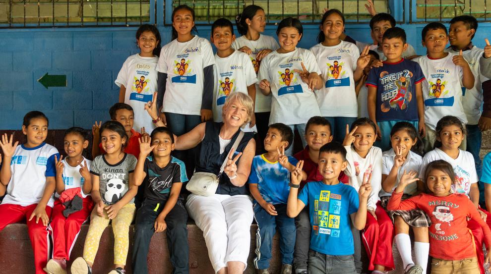 A group of about 20 smiling children holding their hands up and making peace signs, posing with a woman with blonde hair.