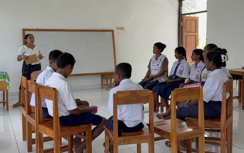 A teacher stands at the front of a classroom, holding a notebook beside a whiteboard, while students sit in a circle participating in discussion.