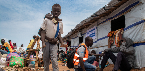 A boy carries a smaller child on his back, walking past a Plan-established temporary shelter at the Sudan border for people fleeing conflict.