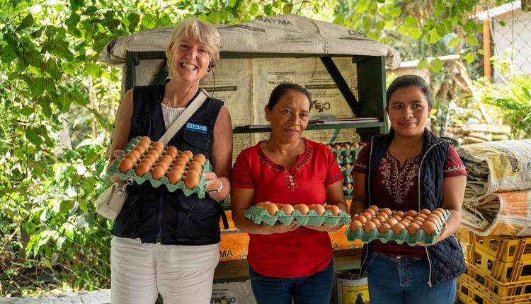 Three women stand together holding crates of eggs.