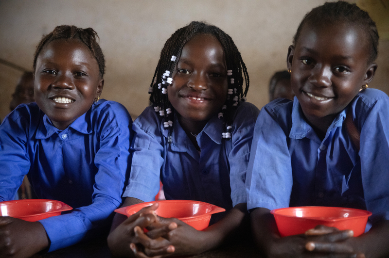 Three children wearing blue shirts standing shoulder to shoulder, each clutching a red bowl.