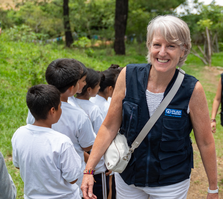 A woman with short blonde hair wearing a dark blue vest smiles with a group of children in white tshirts standing next to her in a line, with their backs to her.