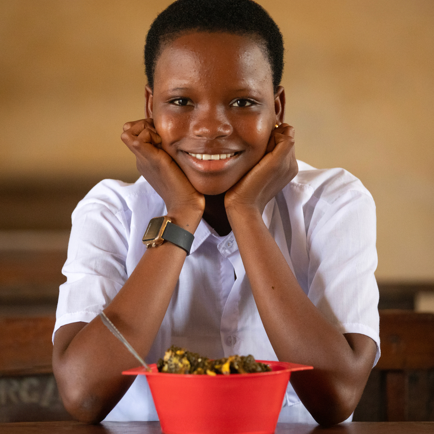 A child wearing a white shirt is sitting at a table smiling with her hands on her chin in front of a red bowl of food.