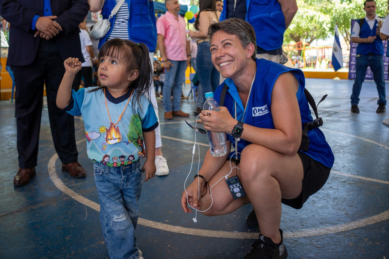 A woman in a blue jacket with the Plan logo kneels beside a young girl.