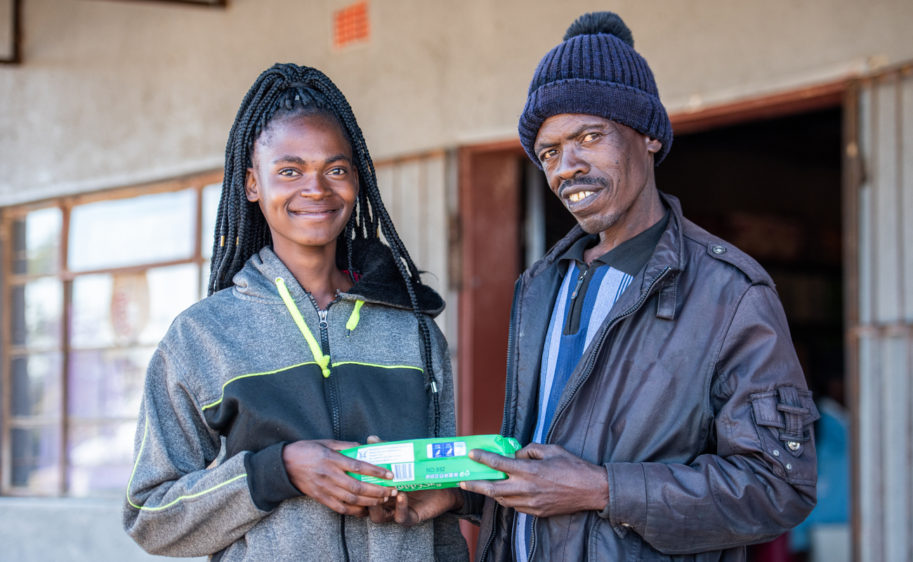 A girl and a man smile and pose for the camera while both holding a box of menstrual pads.