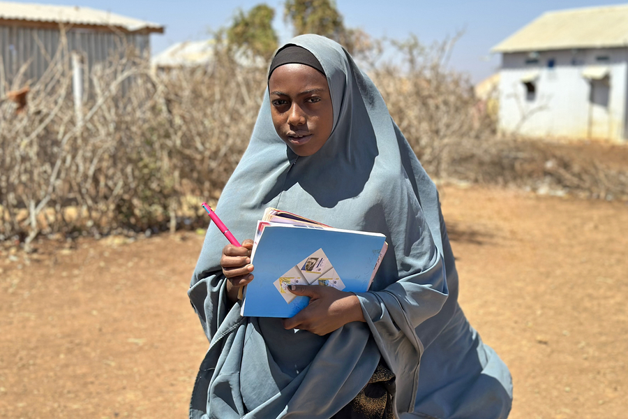 A young girl in a gray hijab holds notebooks and a pen, standing in a dry, outdoor setting with sparse vegetation and buildings in the background.