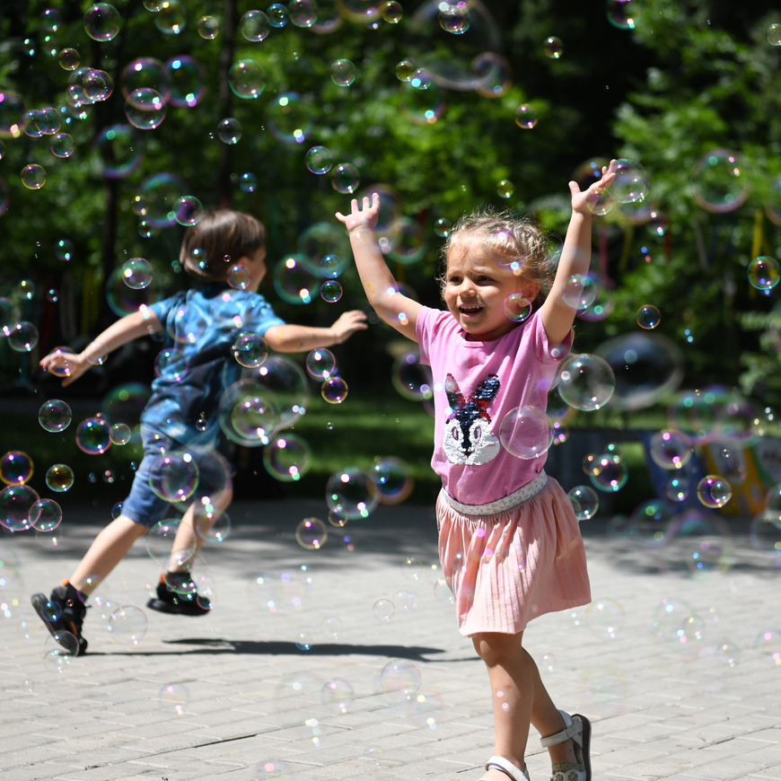 A young girl in a pink shirt and skirt runs through bubbles outside. A boy dressed in blue can be seen in the background.