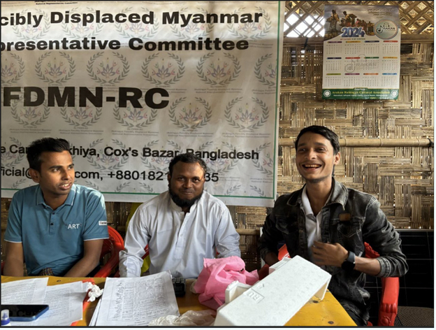 Three men talking and smiling with each other while they’re sitting at a desk.