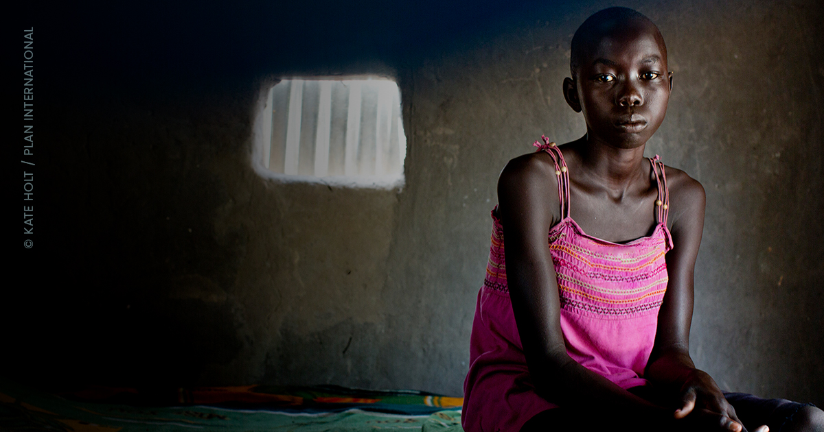 A young girl in a pink dress has a concerned expression as she sits in a dark room in South Sudan