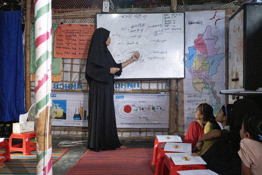A teacher wearing black pointing to writing on a white board while 3 girls look on.