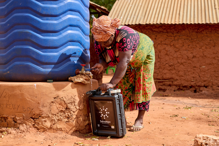 A woman is bent over beside a large water container while she fills up a smaller plastic container that is solar powered and will heat the water. 