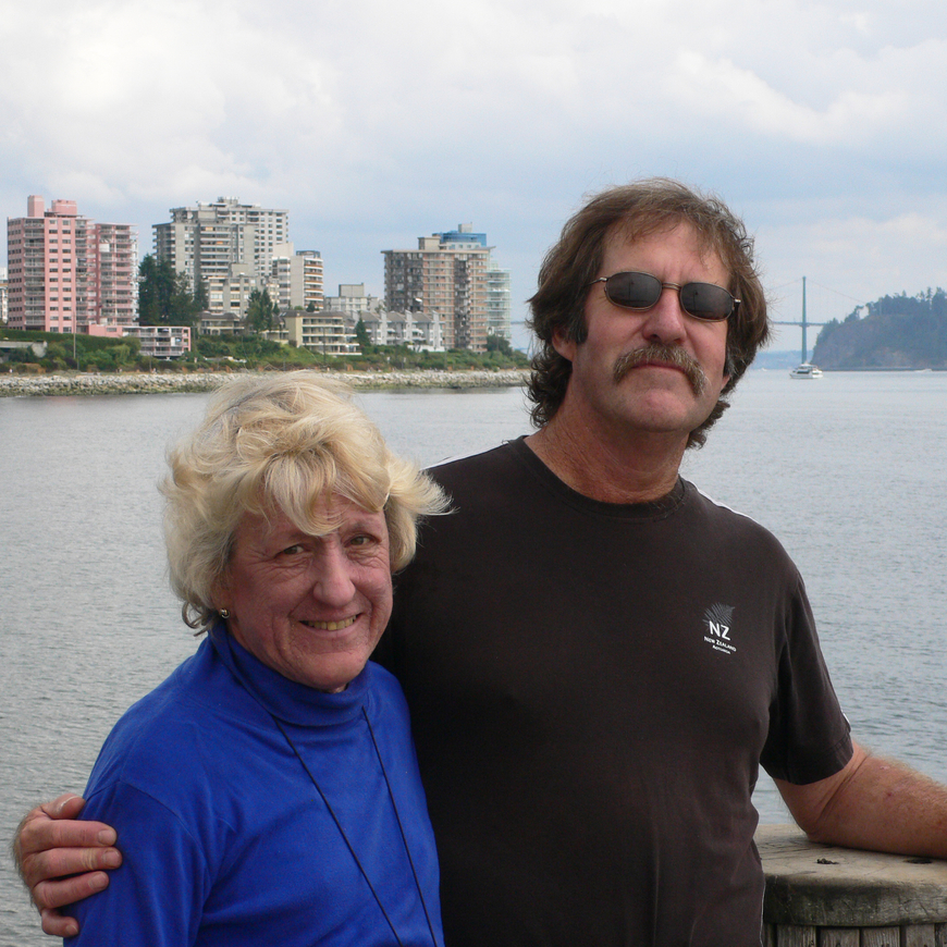A woman in a blue turtleneck stands next to a man in a black t-shirt with sunglasses in front of a waterfront with buildings and a bridge in the background.