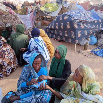 A group of three women surrounded by makeshift tents in a sandy area.