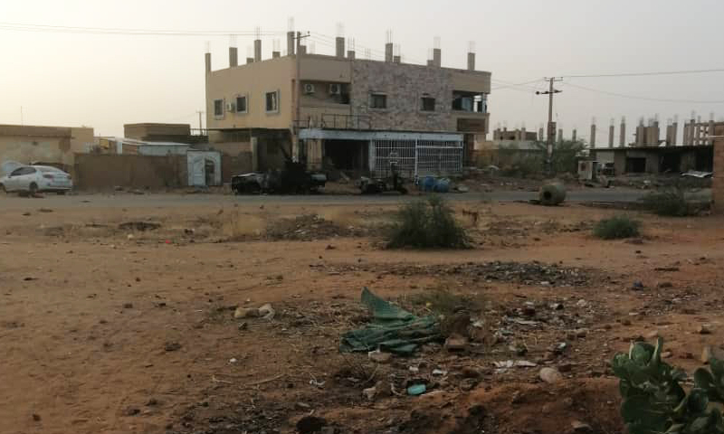 A barren, sandy lot with scattered debris and a few buildings in the background in Sudan.