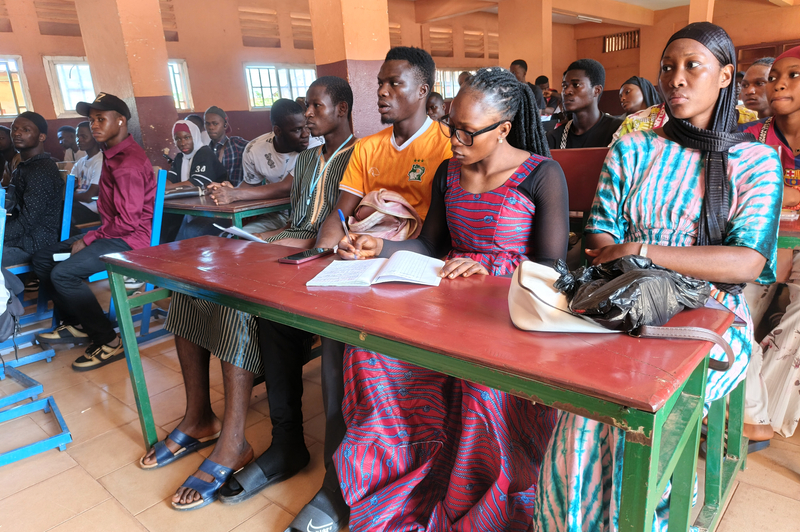 University students sit at desks in a classroom. Some are writing in notebooks while others are looking ahead at the instructor at the front of the room.