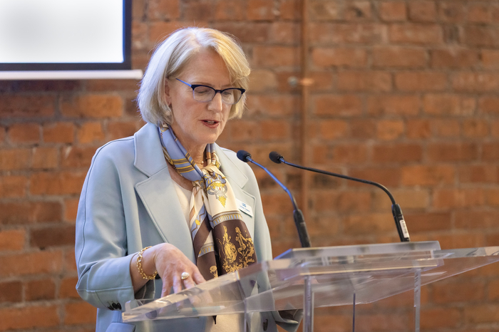 A woman with short blonde hair and glasses speaks at a podium. 