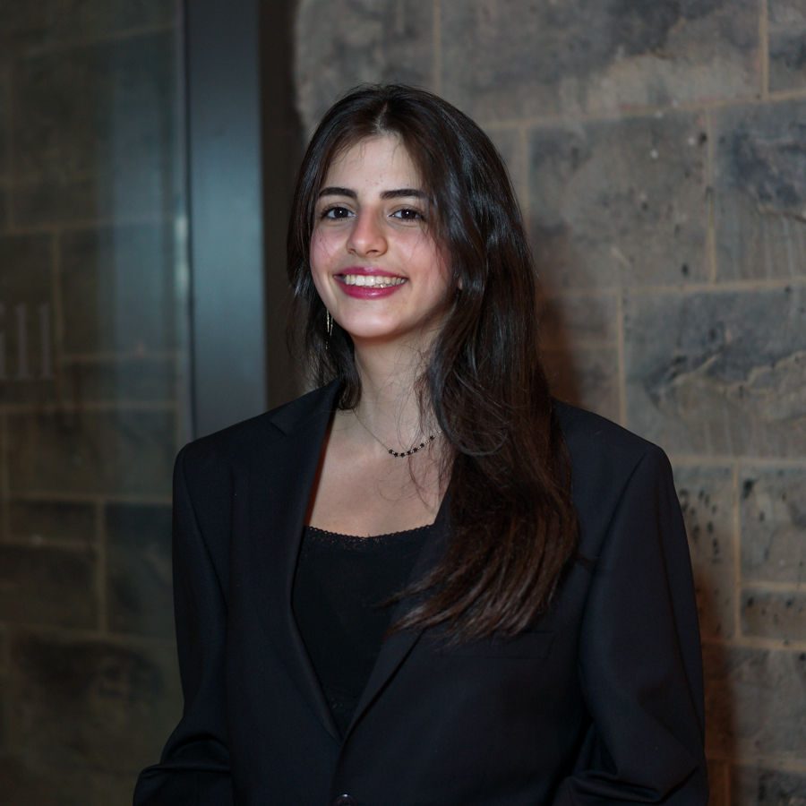 A young woman wearing a black blazer is standing against a brick wall and looking at the camera with her hands clasped. She has shoulder-length dark hair.