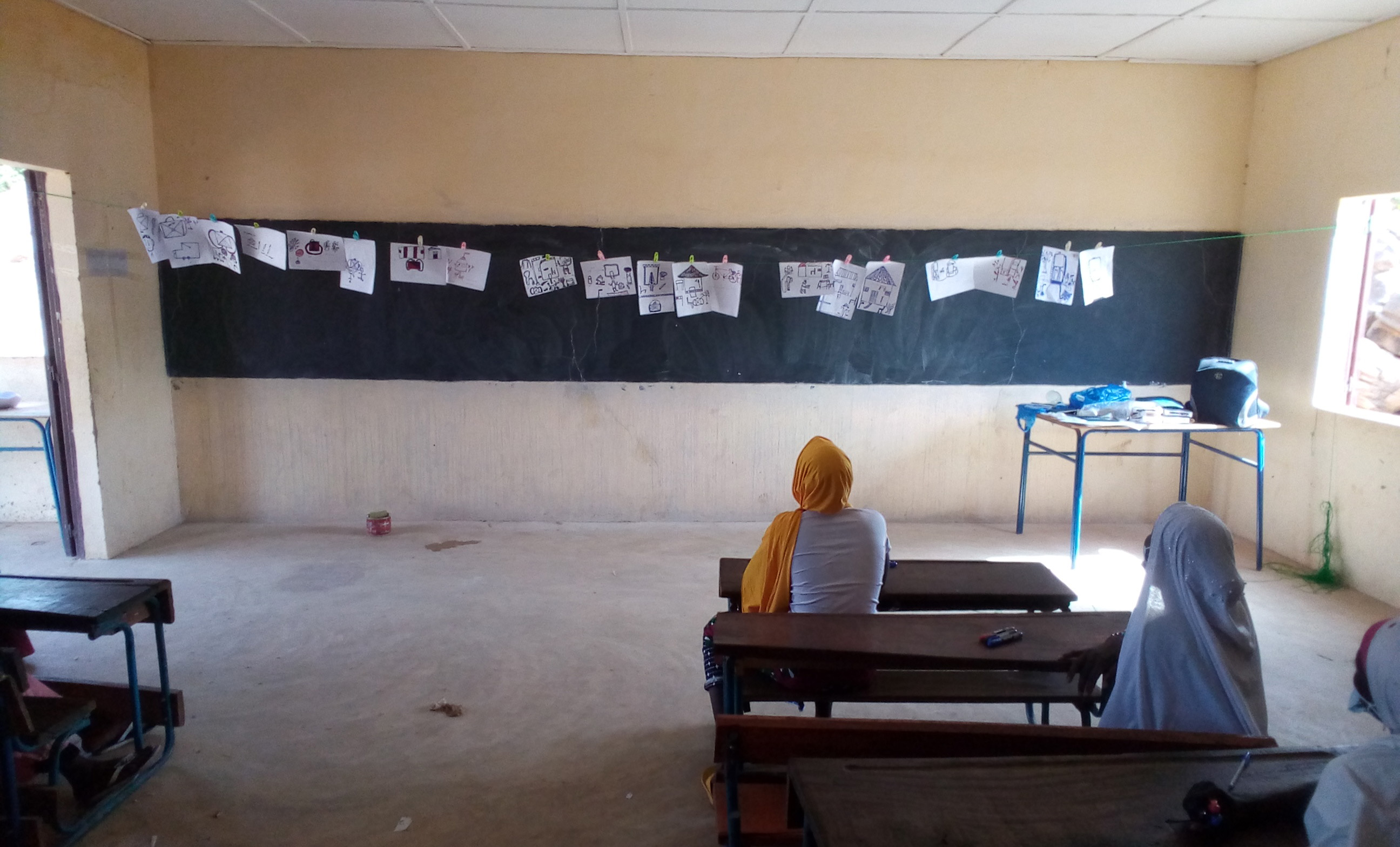 Two girls are sitting in the classroom and looking at drawings hanging in front of the blackboard.