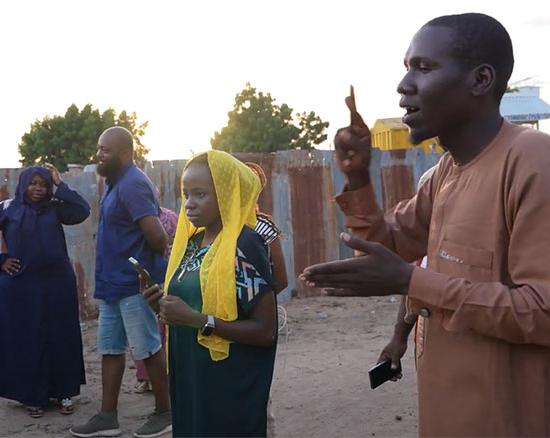 A man signs to a man standing across from him. A woman in between them films using a phone on a tripod. 