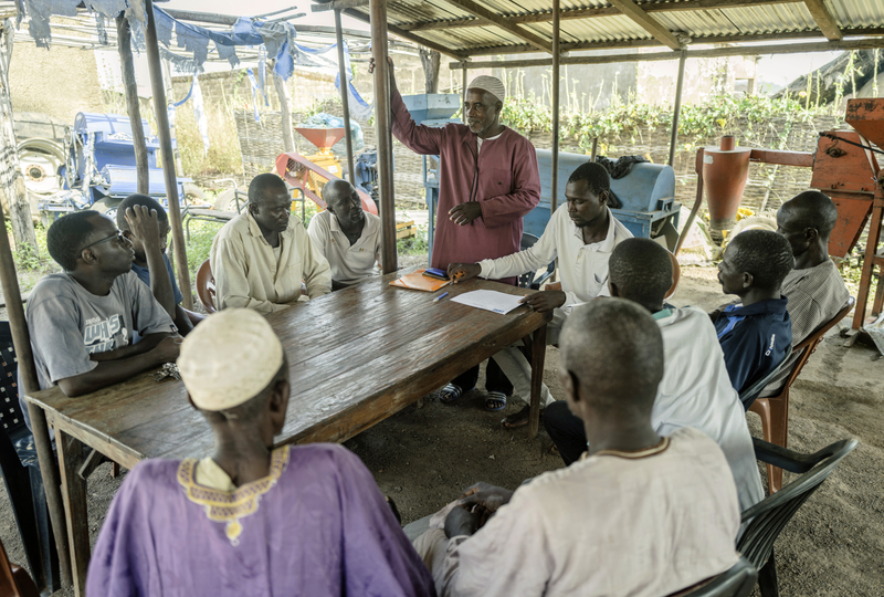 A group of men sitting around a wooden table are listening to a man wearing a red top standing up at the end of a table speaking to the group.