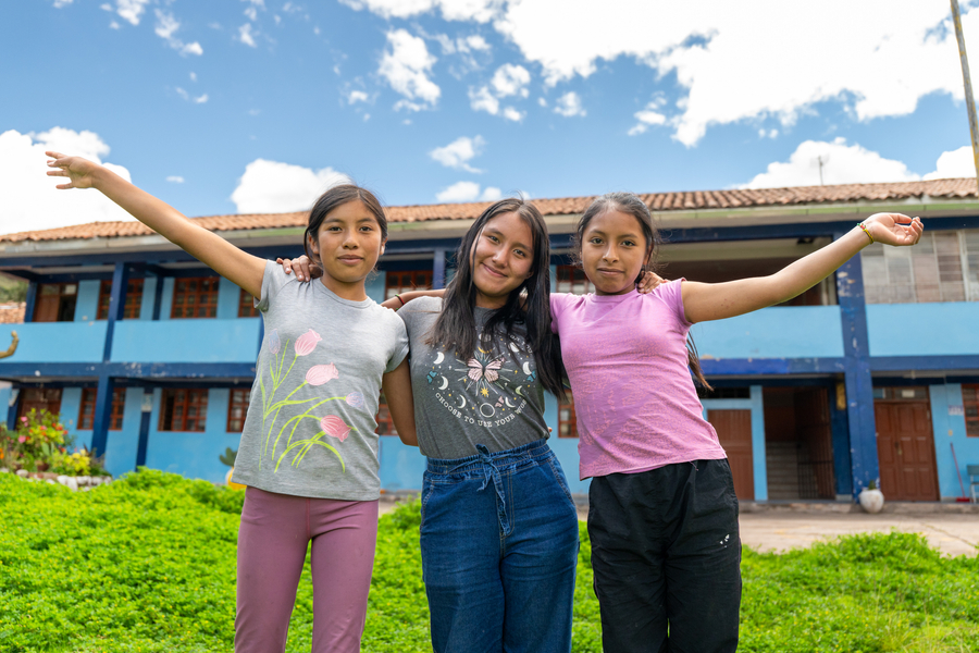 Three girls stand together outside a blue school building, smiling and posing against a backdrop of a clear sky and green grass.