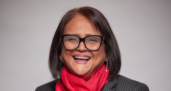 A smiling woman with dark hair and glasses, wearing a black and white pinstripe blazer and a bright red scarf, stands facing the camera against a plain grey background.