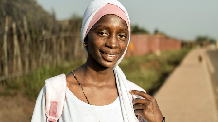 A smiling teenage girl wearing a white and pink head scarf and a white t-shirt stands on the sidewalk beside a road in rural Senegal.