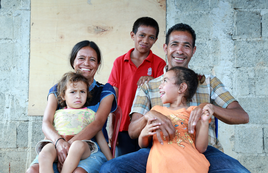 A smiling family poses together, with two adults and three children, showcasing a warm and joyful atmosphere against a simple background.