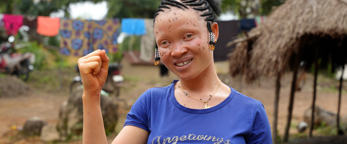 A smiling young woman with braided hair raises her fist confidently, standing outdoors with colorful clothes hanging in the background.
