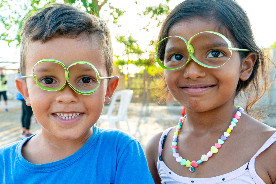 Two children smile at the camera, wearing oversized green paper glasses.