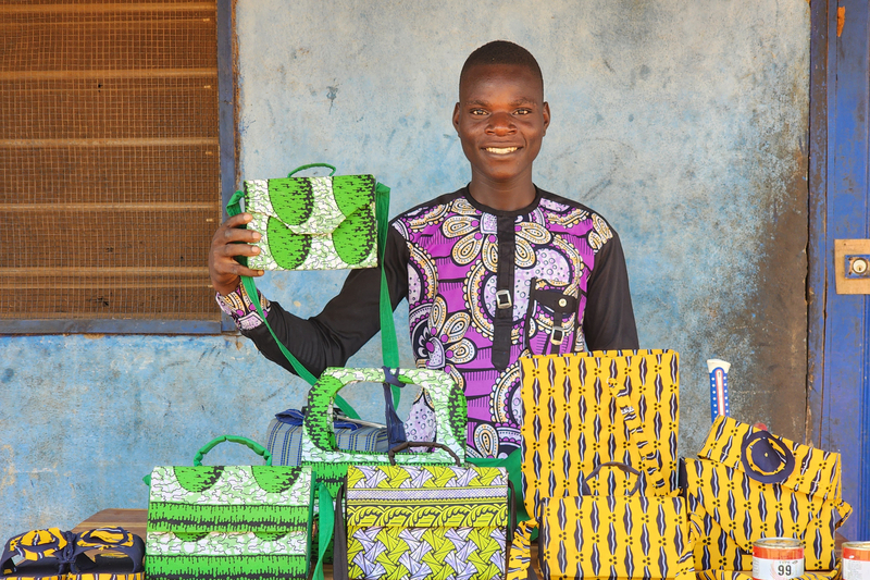 A young man holds up a green handbag he made while other colourful handbags are displayed in from of him on a table.