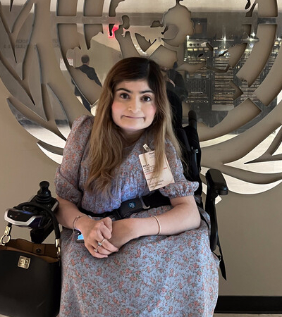 A woman with long light brown hair wearing a floral dress sits in a powered wheelchair.