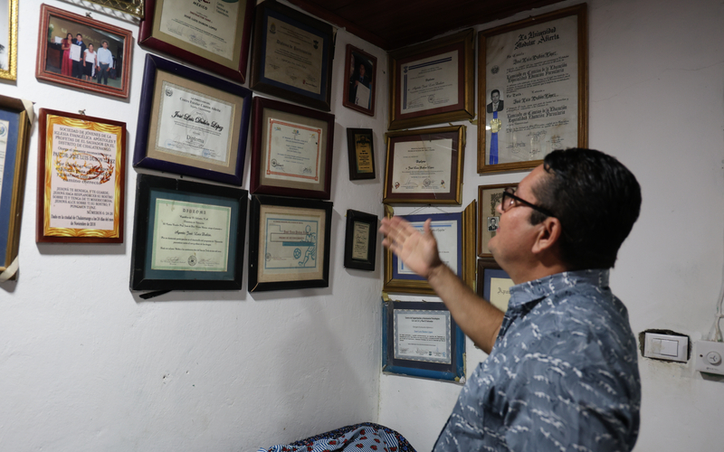A man wearing glasses and a patterned shirt stands in a room, pointing at a wall filled with framed certificates, awards, and photographs.