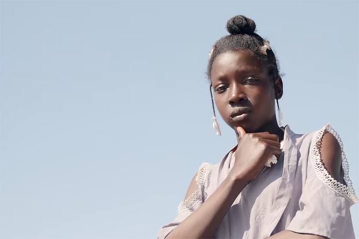 A teen girl in Haiti who lives in a displacement camp, looking directly at the camera, with a video “play” button overlaid. She is one of three girls featured in our mini-documentary on the ongoing crisis in Haiti.