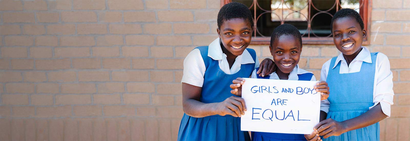 Three girls grinning and holding a "Girls and boys are equal" sign, while standing shoulder by shoulder in front of a school