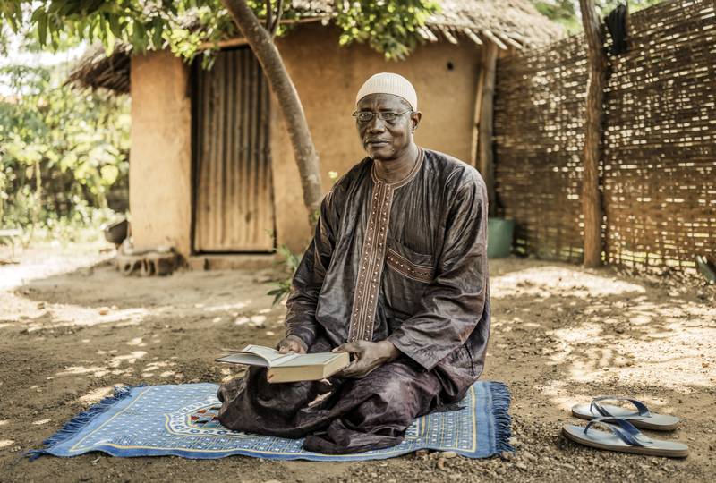 An older man in traditional Islamic dress is holding a book while sitting on a rug on the ground outside.