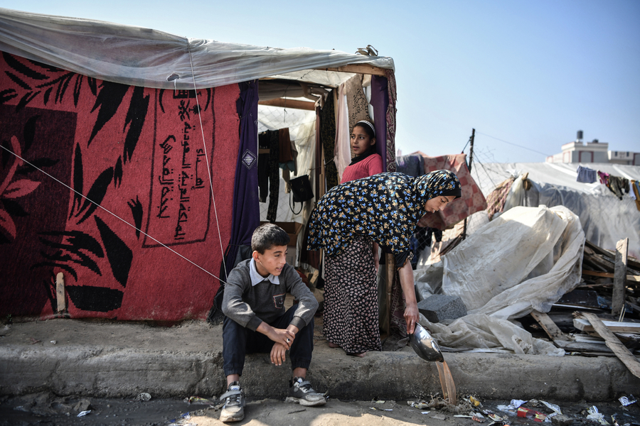 A boy sits outside a makeshift shelter while a woman cleans nearby, surrounded by a temporary settlement. 