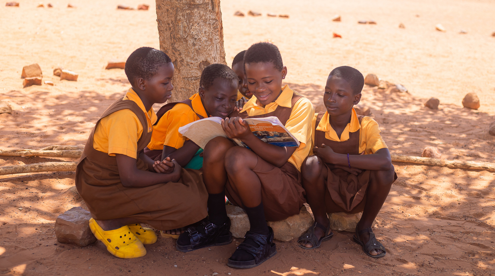 Four children sitting under a tree in a dry, open area, reading a book together. Children are wearing matching yellow shirts and brown uniforms, with one child wearing bright yellow shoes.